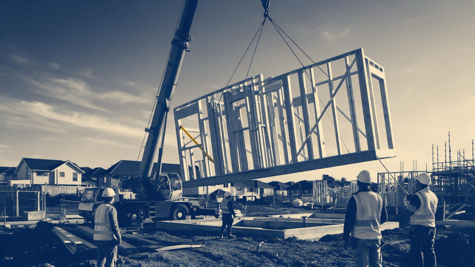 A crane lifting a prefabricated timber house frame into place on a new housing estate, workers watching from below in the morning light