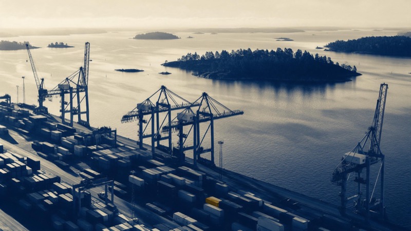 A Finnish seaport seen from above in early morning light, cranes and stacked containers along the quay, pine-covered islets offshore