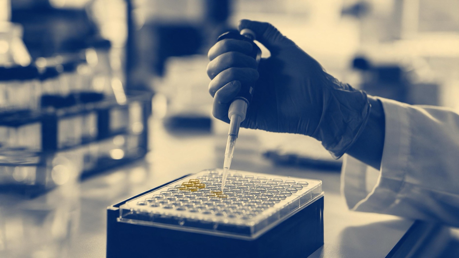 A researcher's gloved hand holding a pipette over a tray of tiny sample wells, shot close in bright lab light