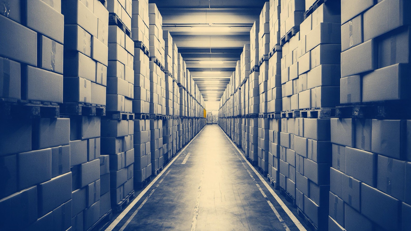 A warehouse aisle of identical plain cardboard boxes on pallets stacked to the ceiling, lit by flat fluorescent light and lined with floor markings