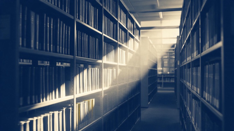 Long aisles of tall steel shelving in a university research library, bound journals with plain spines, dust turning in a shaft of afternoon light