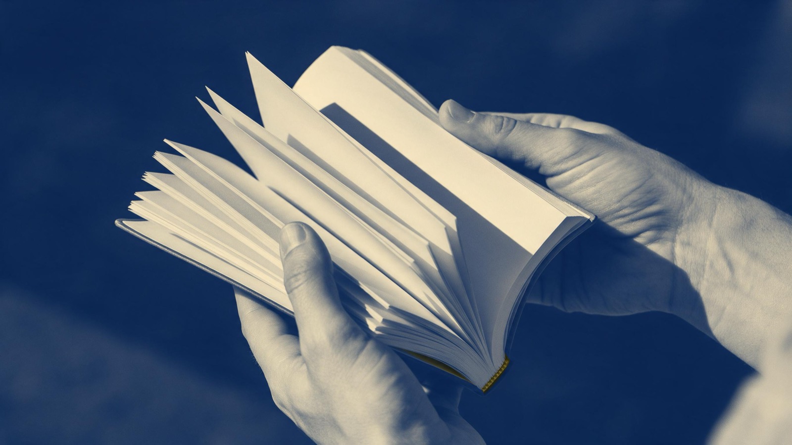 A scholar's hands opening a freshly printed book under a reading lamp, crisp pages fanning, only the hands and the pages in the frame