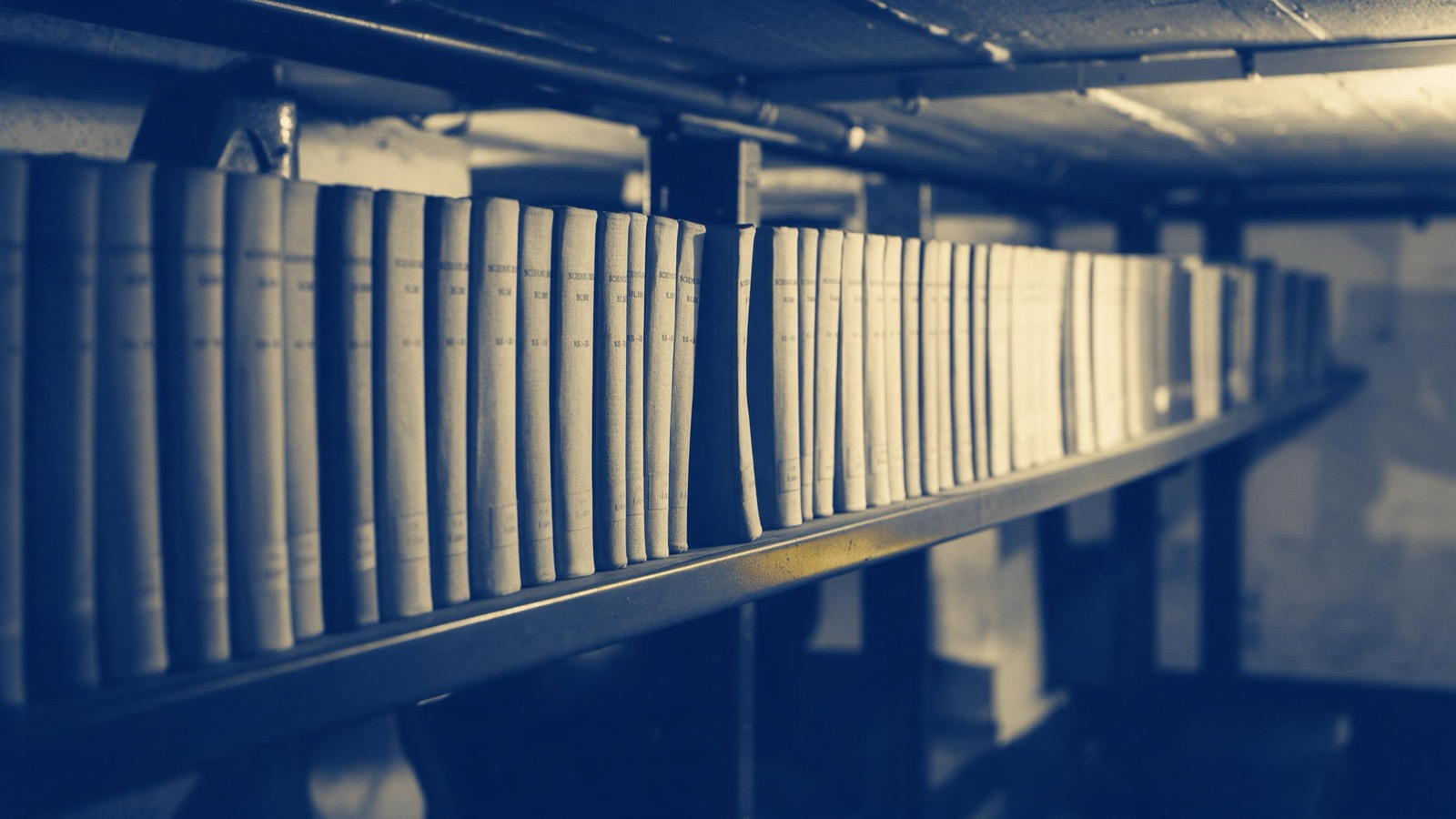 Bound volumes of old scientific journals on a long steel shelf in a basement archive, plain cloth spines, one gap where a volume is missing