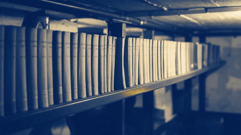 Bound volumes of old scientific journals on a long steel shelf in a basement archive, plain cloth spines, one gap where a volume is missing