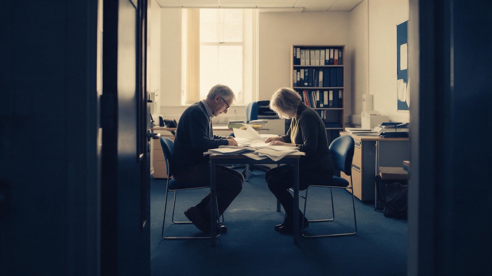 Two academics seated across a table in a quiet university office, seen from a doorway at a distance, heads bowed over a pile of papers