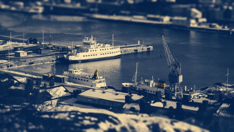 A Nordic harbour seen from a hill in winter, passenger ferries moored along granite quays beside low rooftops and cranes, thin ice at the water's edge