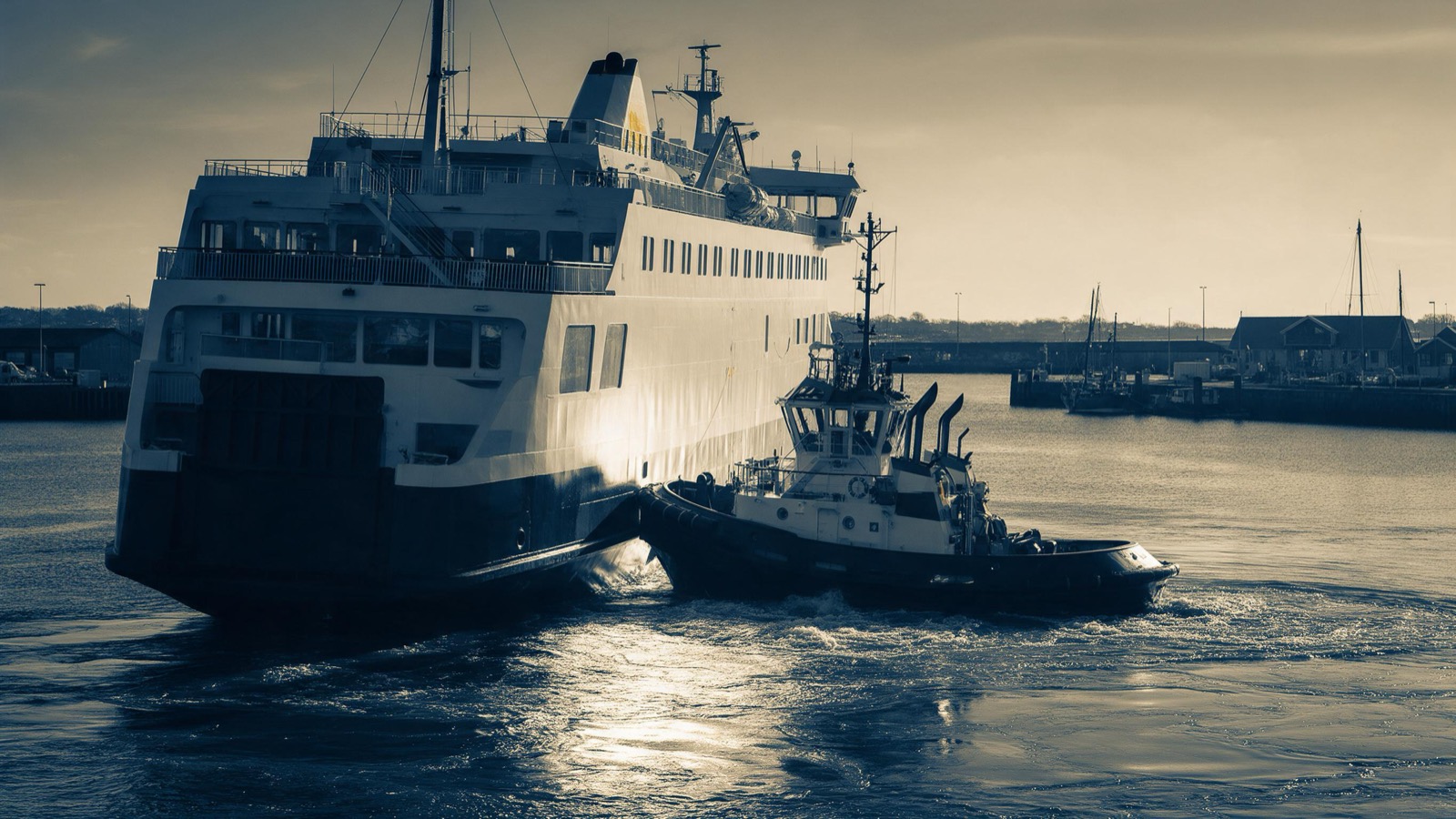 A tug pushing against the flank of a large ferry as it swings in the harbour basin, low winter sun glinting off the churned water