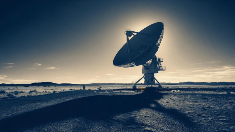 A large radio antenna dish tilted toward a low sun on a high desert plain, its long shadow stretching across the ground