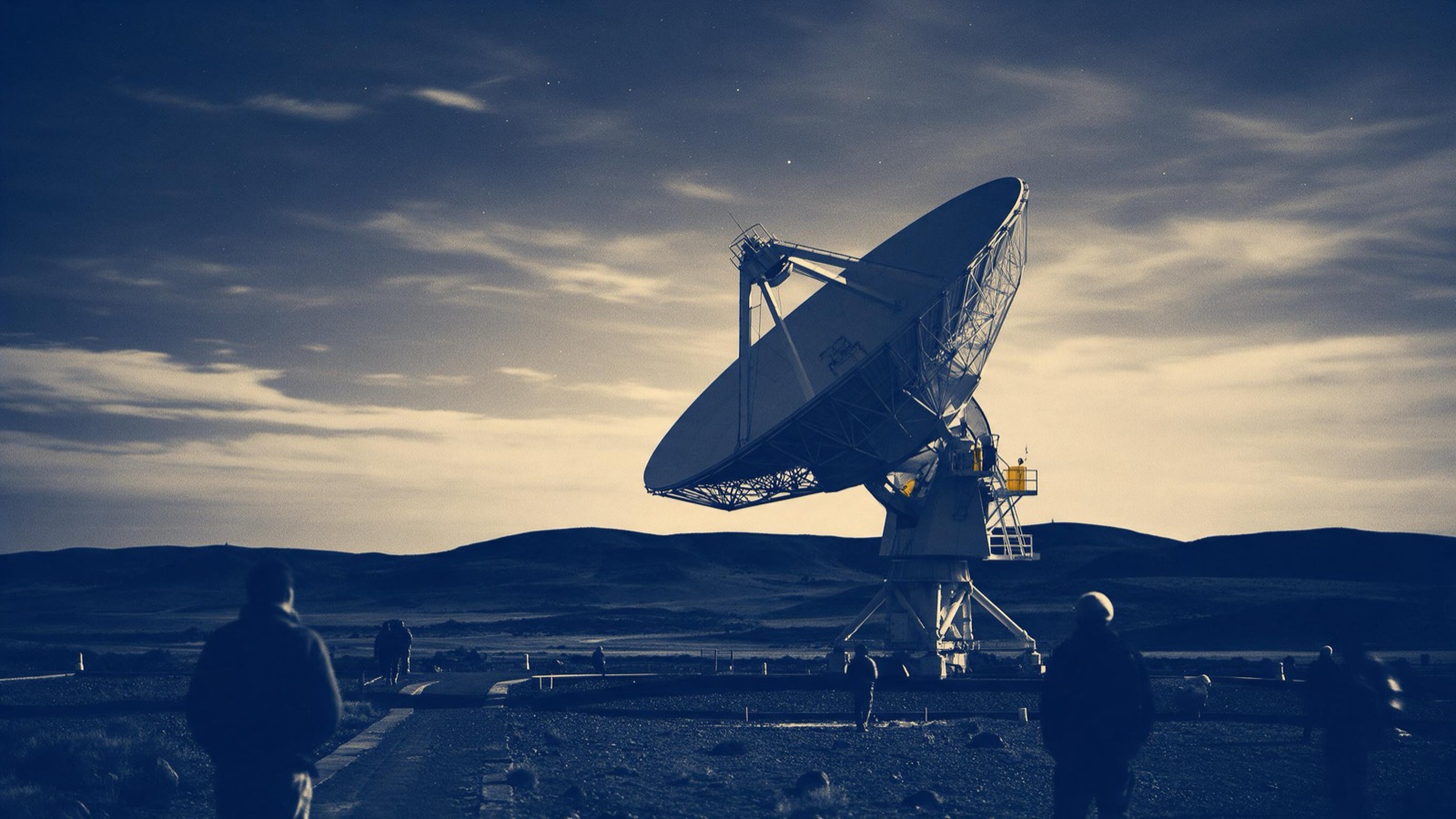 Technicians seen from behind climbing a service ladder on the steel frame of a big antenna dish, the sky pale behind them