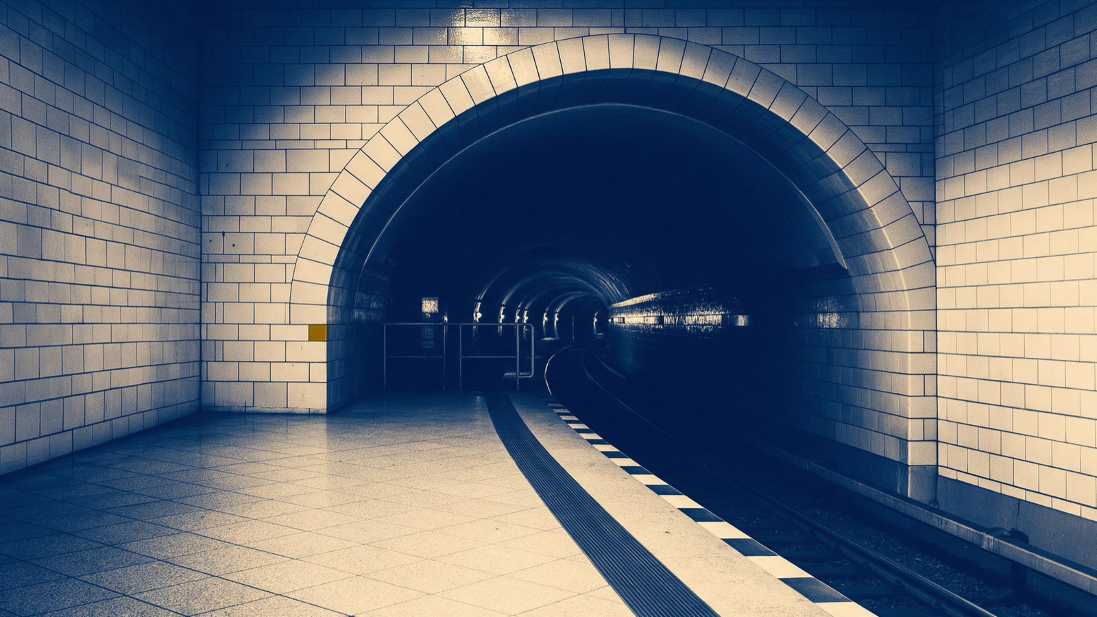 A deep tiled underground station with a curved arch, empty platform and tracks receding into the dark of the tunnel, lit in warm light
