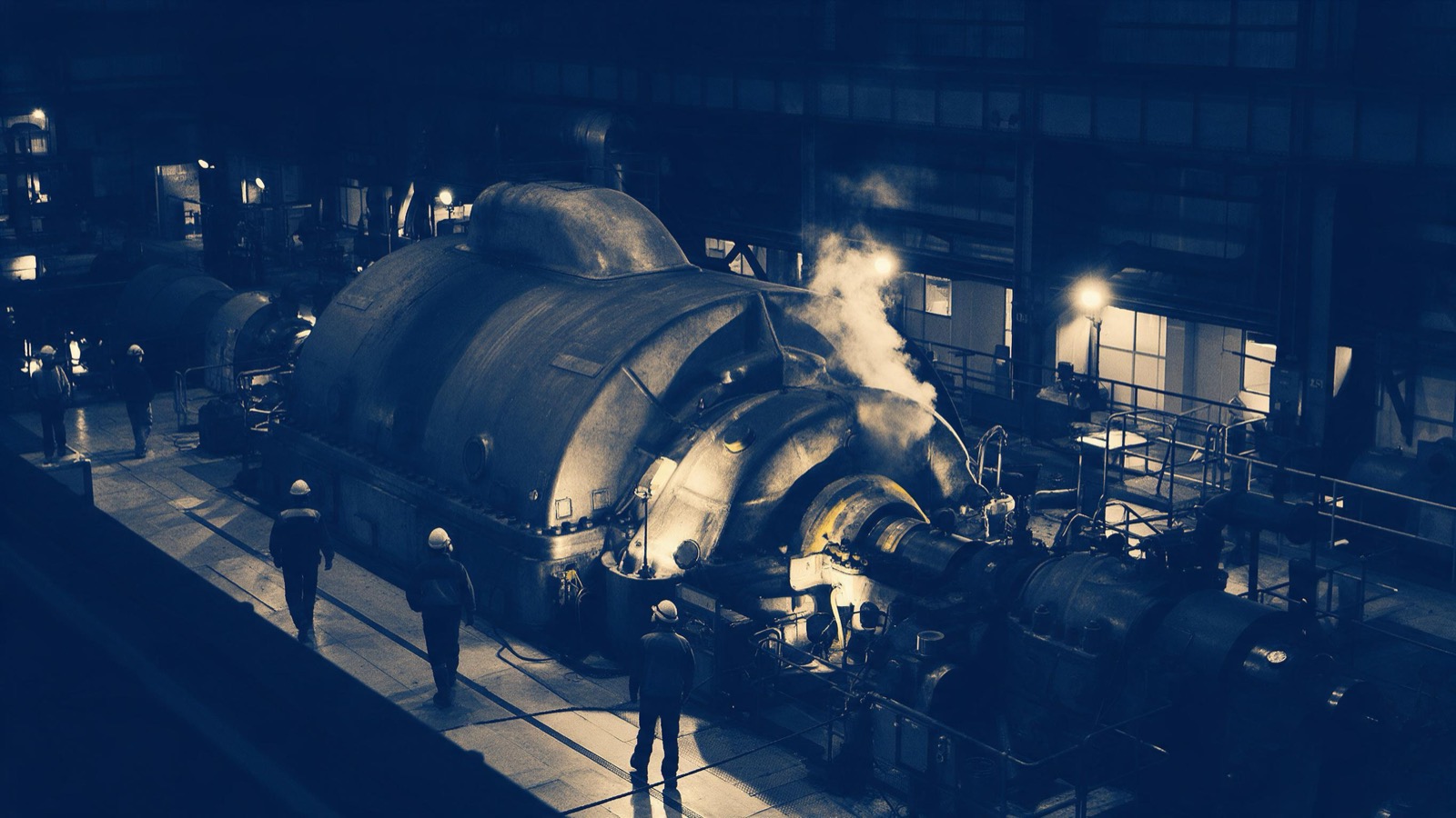 Engineers in hard hats and hi-vis jackets seen from behind walking along a gallery inside a turbine hall beside a huge generator casing