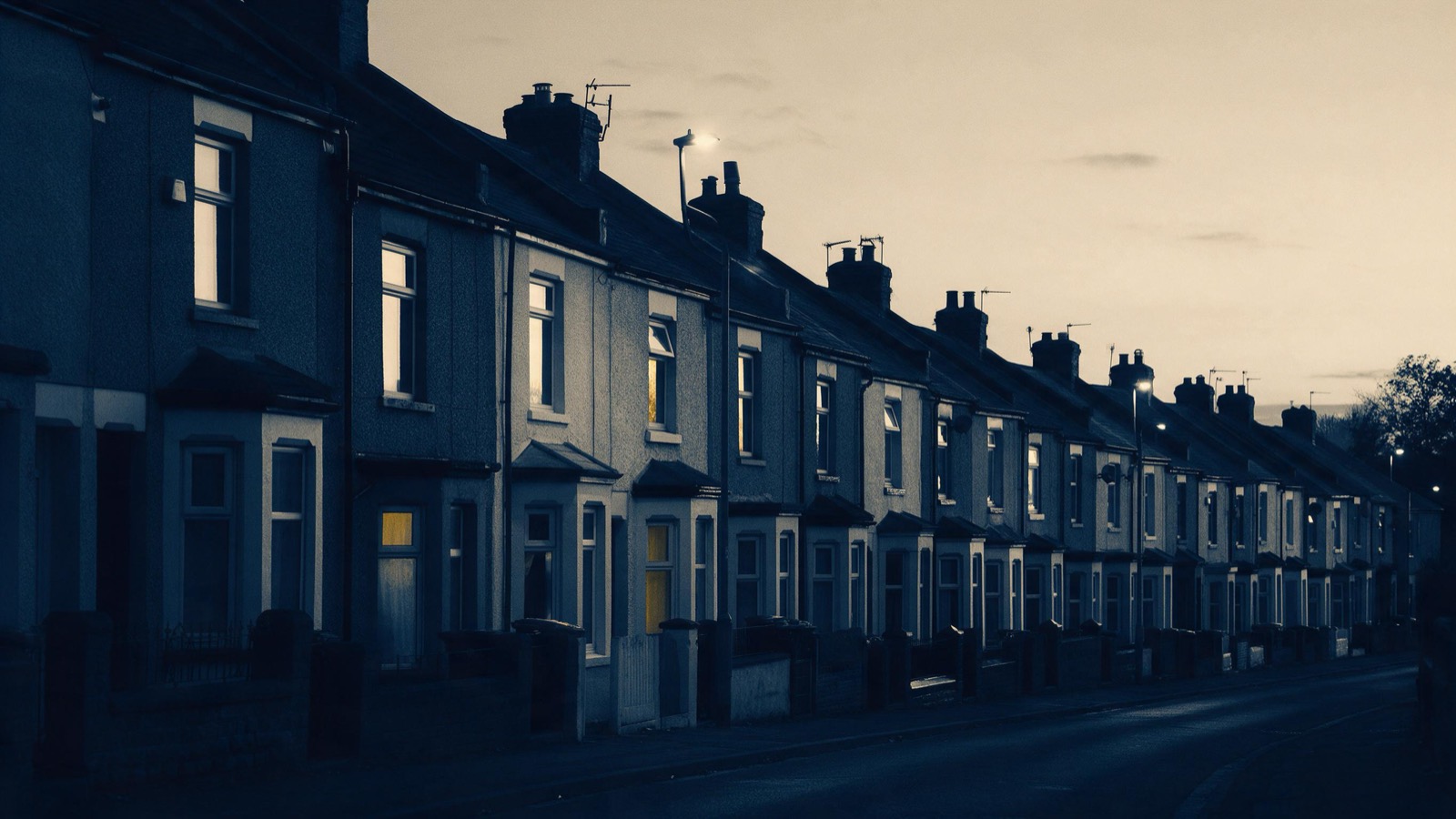 Street lamps flickering on one after another along a terraced British street at dusk as the windows of the houses begin to glow yellow