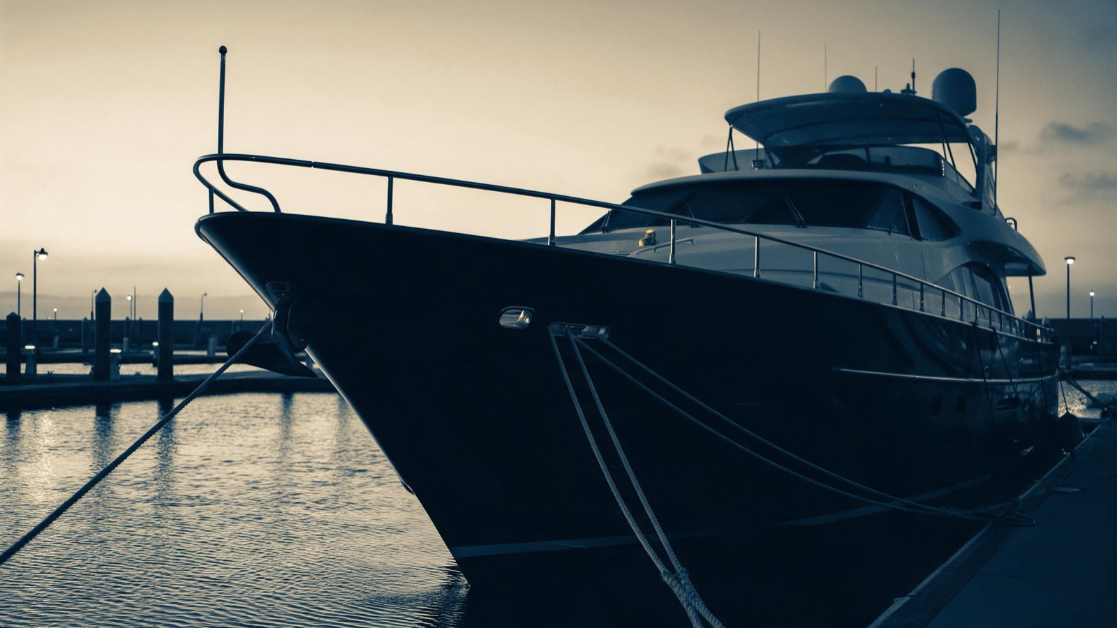 A large private yacht moored alone and dark at an empty marina at dawn, ropes taut, dockside lamps still burning, no one aboard