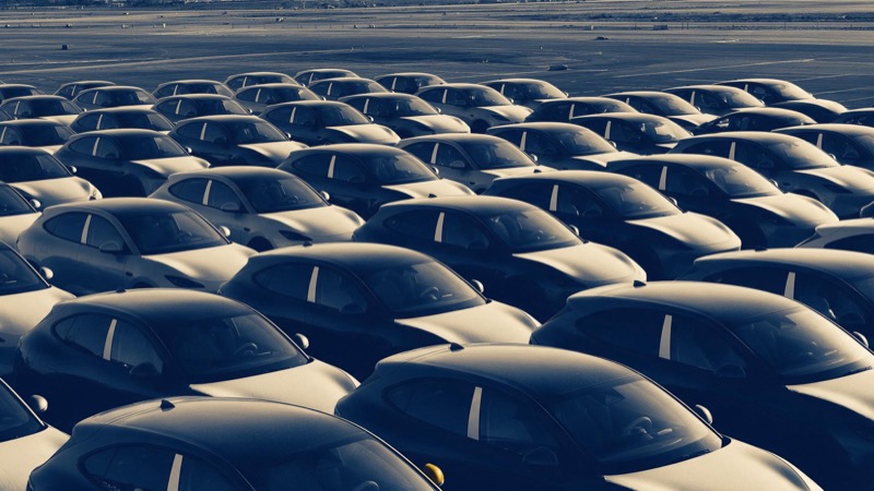 Dozens of identical unbadged new cars parked in tight rows on a vast airfield lot, seen from above in low sun