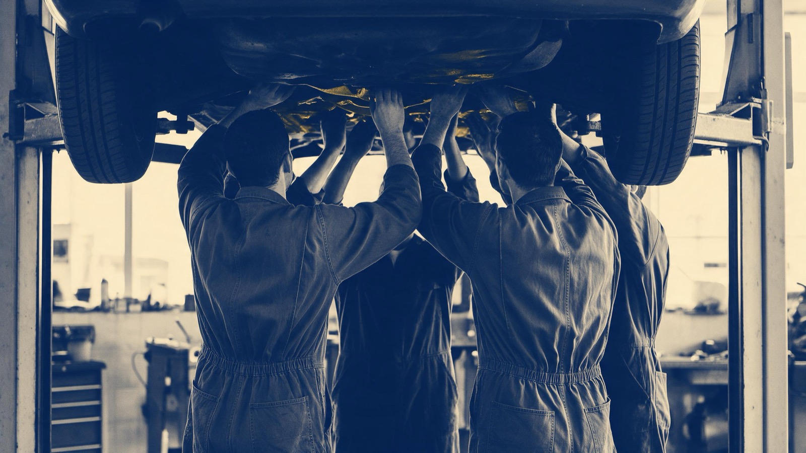 Mechanics in overalls seen from behind under a car raised on a hydraulic lift in a bright workshop, hands reaching up to the underside
