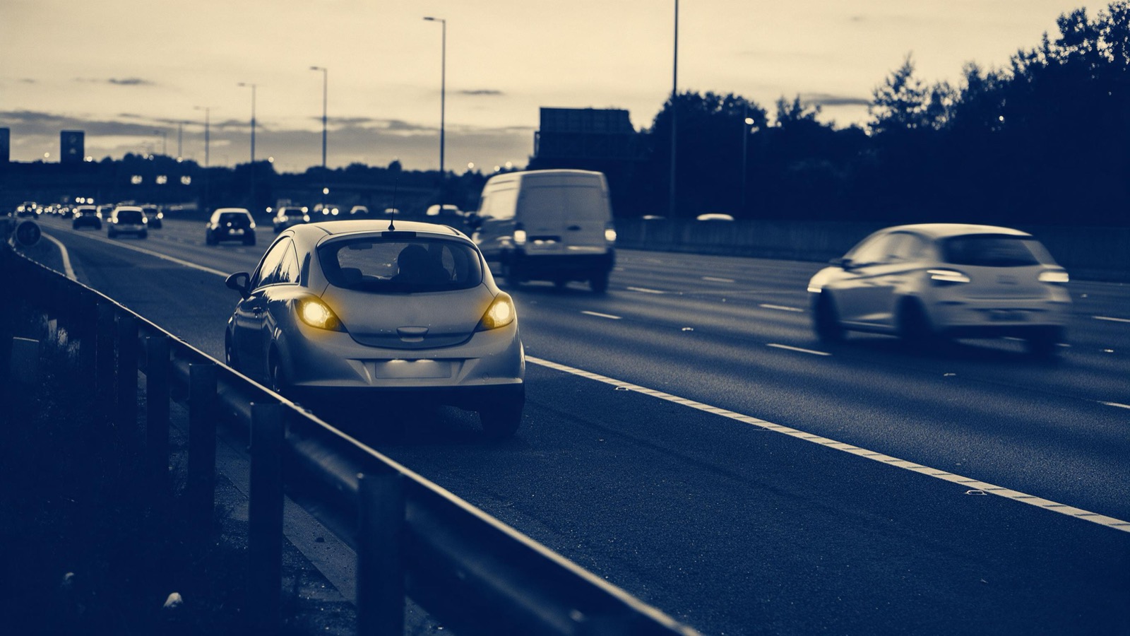 A car pulling onto the hard shoulder of a motorway at dusk with its hazard lights flashing, other traffic streaming past