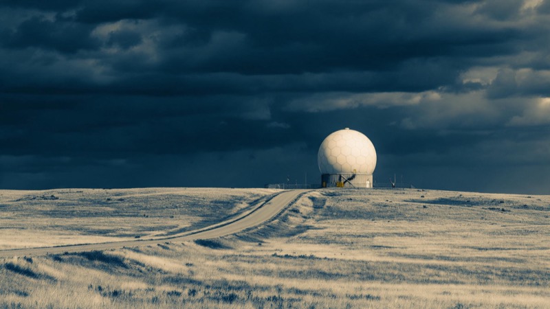 A white weather radar dome on a plains hilltop under heavy dark cloud, a lone service road winding up toward it