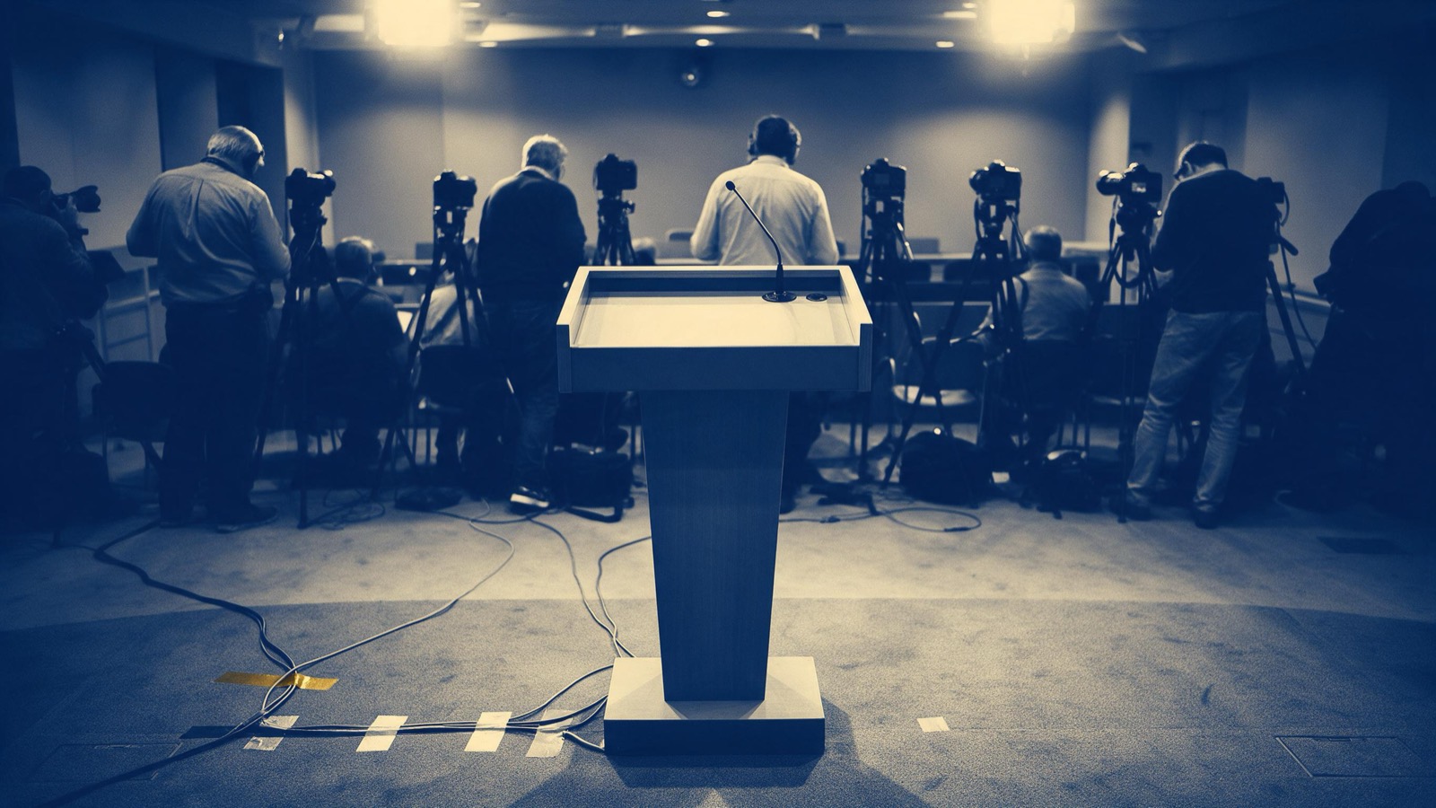 A lone speaker seen from behind stepping up to a podium ringed with microphones before a hushed press room, camera flashes catching the front rows